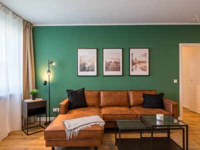 Wohnzimmer mit grüner Akzentwand, braunem Sofa und stilvollen Bildern – modernes Serviced Apartment